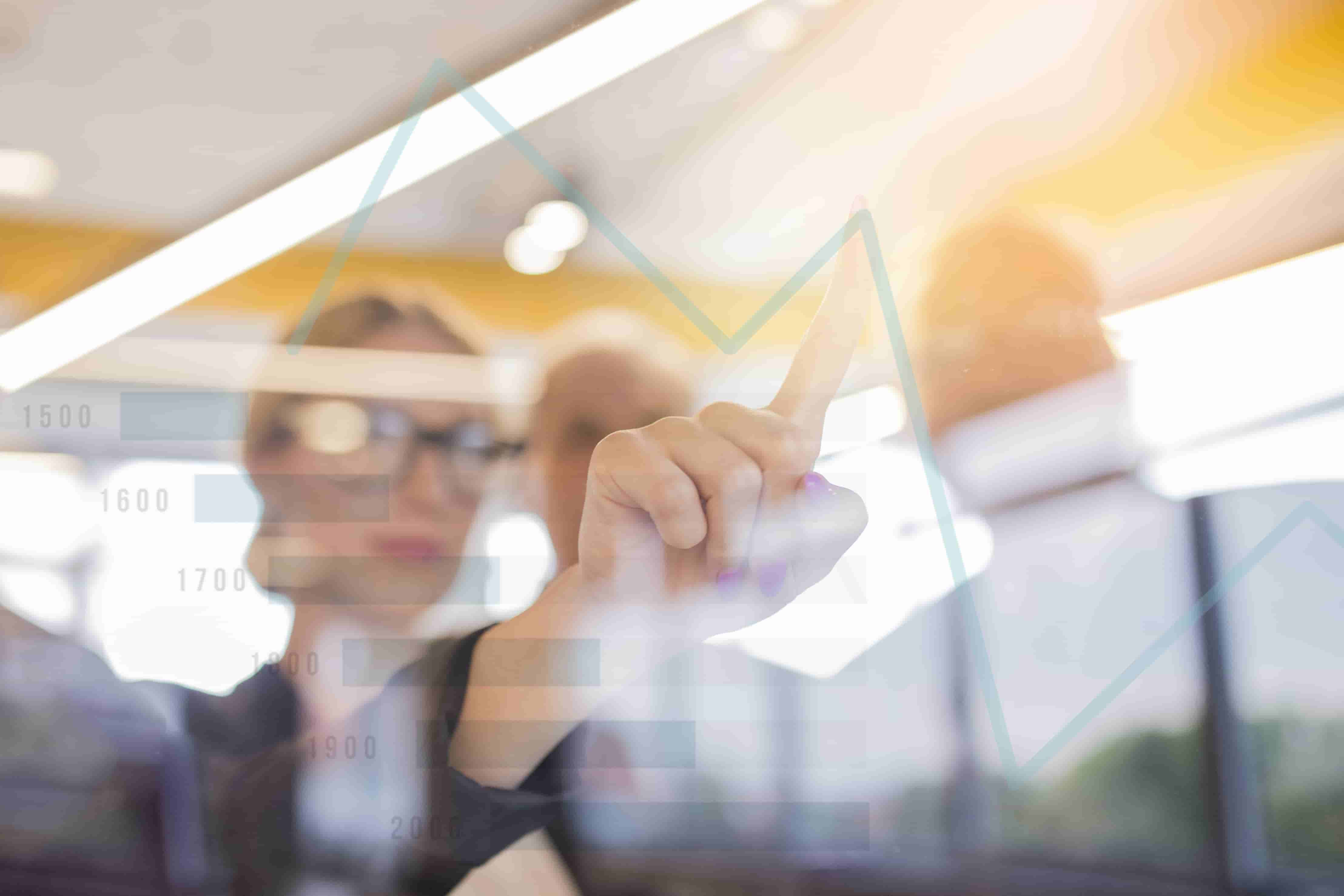 A person wearing glasses points to an ascending graph line on a transparent screen. Multiple bar charts with numerical labels suggest data analysis or financial growth driven by nearsourcing. The background shows other indistinct people in a well-lit office environment, discussing hiring solutions. CodersLink 2024.