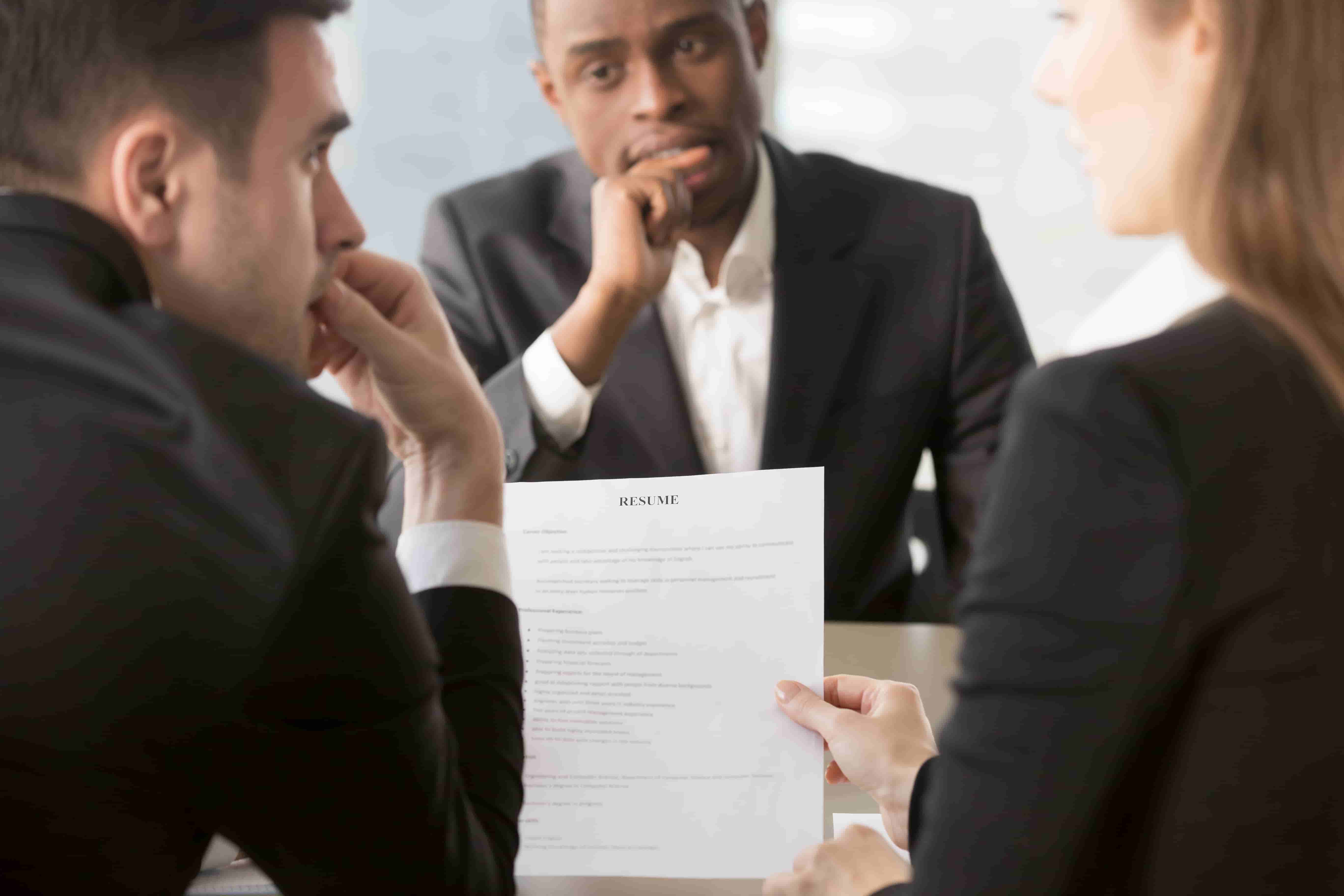 Three individuals in formal business attire sit around a table during a meeting. Two of them are reviewing a resume held by one person. They look focused and engaged in conversation, likely discussing the candidate's qualifications and how nearshoring talent from Latin America could be an optimal hiring solution. CodersLink 2024.