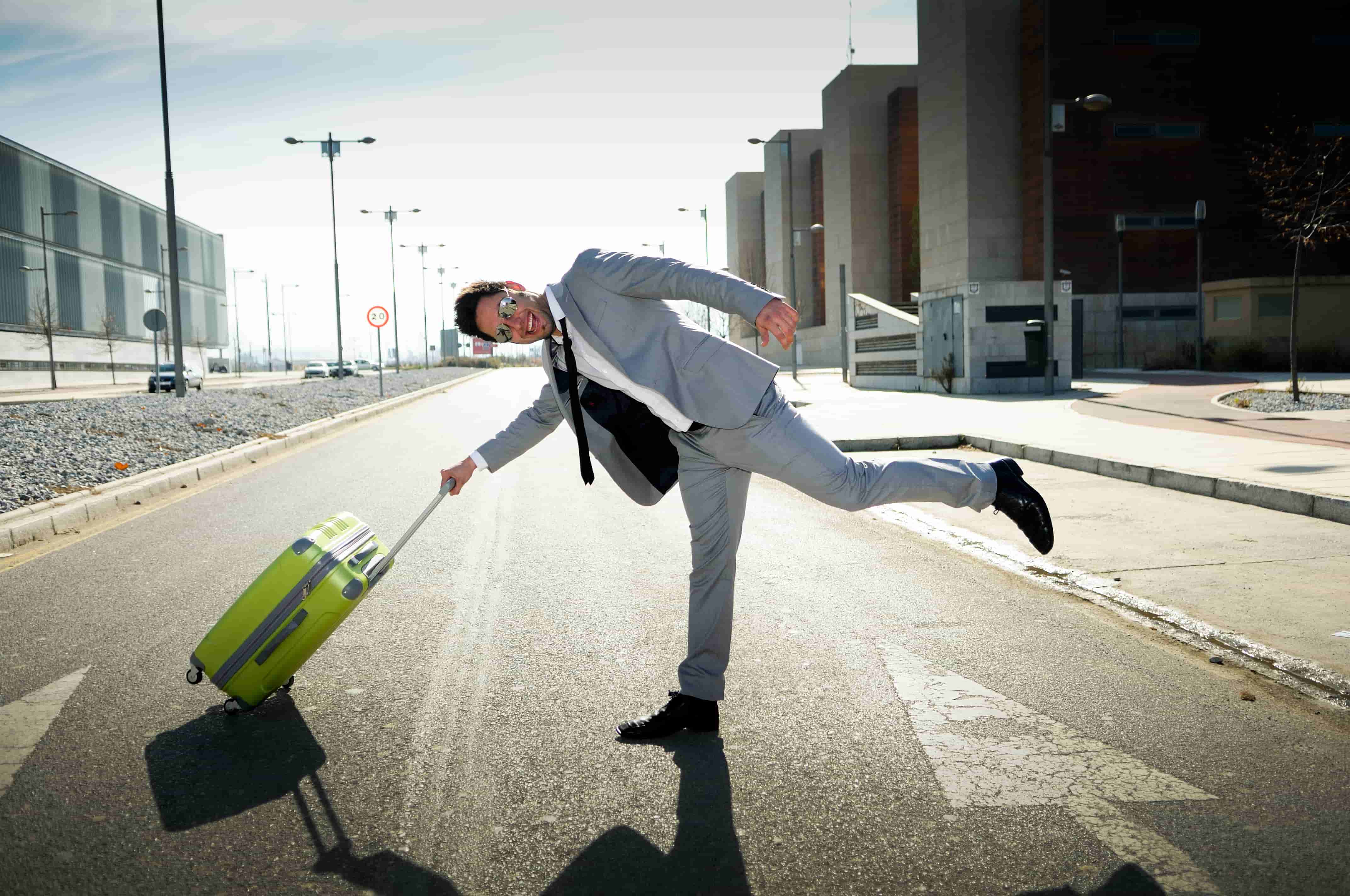 A man in a light gray suit and dark sunglasses playfully balances on one leg while holding a green suitcase on a deserted urban street in Mexico. The street is lined with modern buildings, and the weather appears sunny. CodersLink 2024.