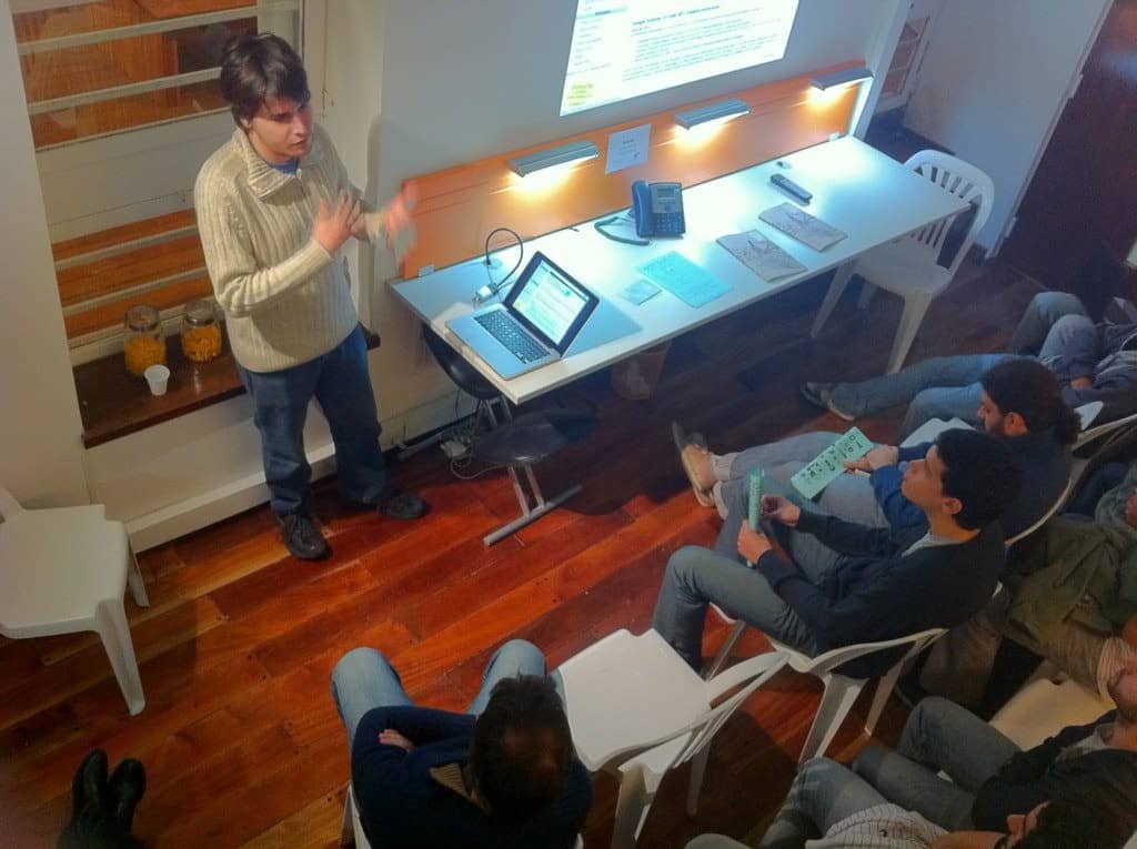 A person stands and gives a presentation on recruitment solutions with a laptop and projected screen in front of a seated audience. The room has wooden floors and white chairs, with various papers and a phone on the table beside the presenter. CodersLink 2024.