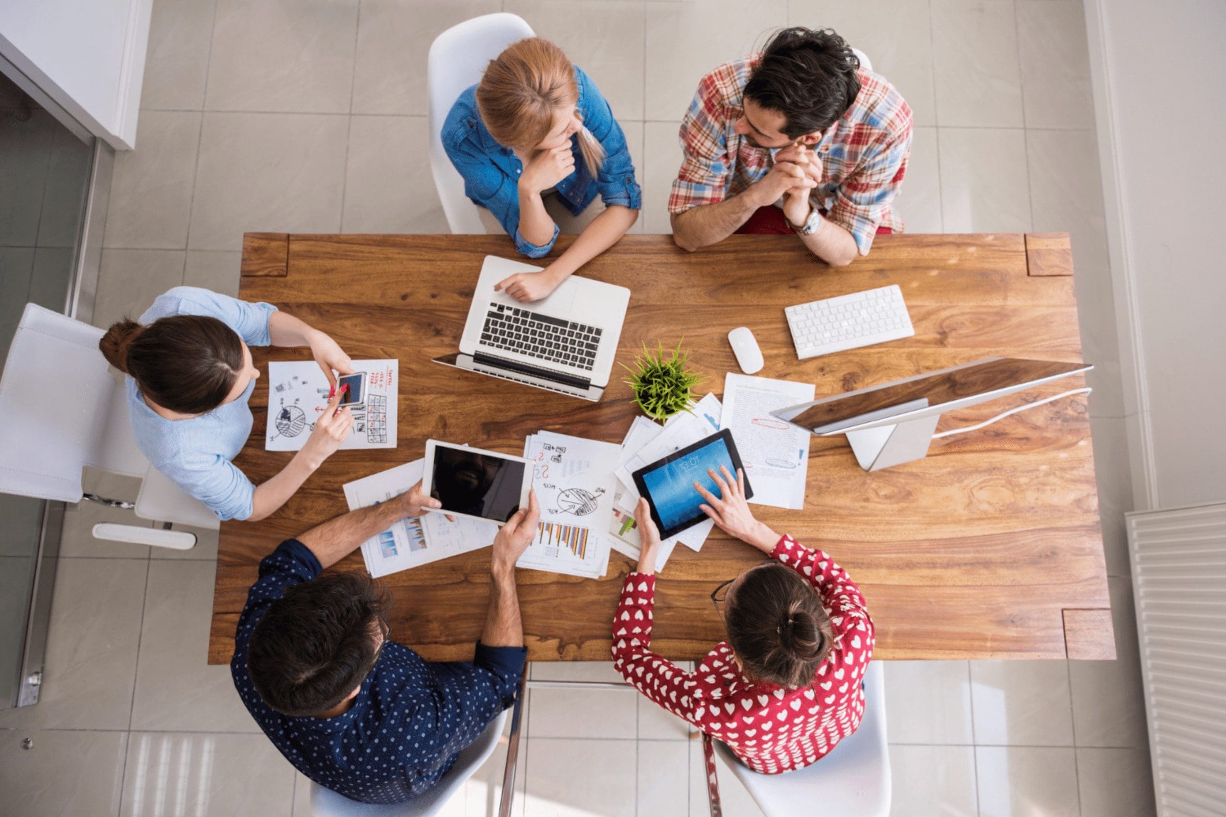 A group of five people sit around a rectangular wooden table, collaborating on various tasks. They have laptops, tablets, documents, and graphs in front of them. They appear to be engaged in a discussion about their employer of record in a modern office setting. CodersLink 2024.