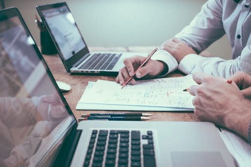 Two people are working at a desk with two laptops. They are reviewing documents and taking notes with pencils. The scene captures a collaborative work environment, with focus on onboarding software engineers. Coffee cups are barely visible in the background. CodersLink 2024.