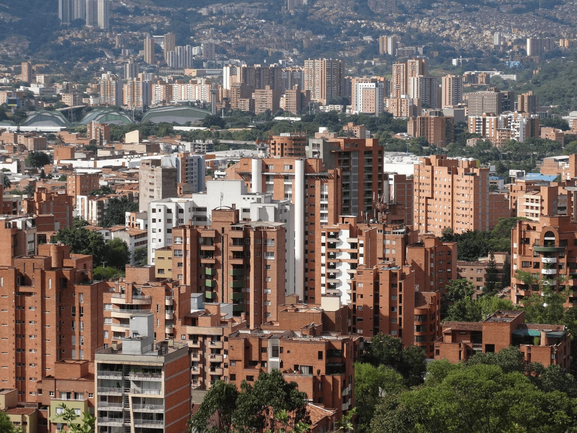 An aerial view of a city features numerous tall, red brick residential buildings in the foreground and a mixed skyline of white and brown high-rises in the background. The landscape, set against densely forested mountains, is as dynamic as an Employer of Record in Colombia navigating diverse employment laws. CodersLink 2024.