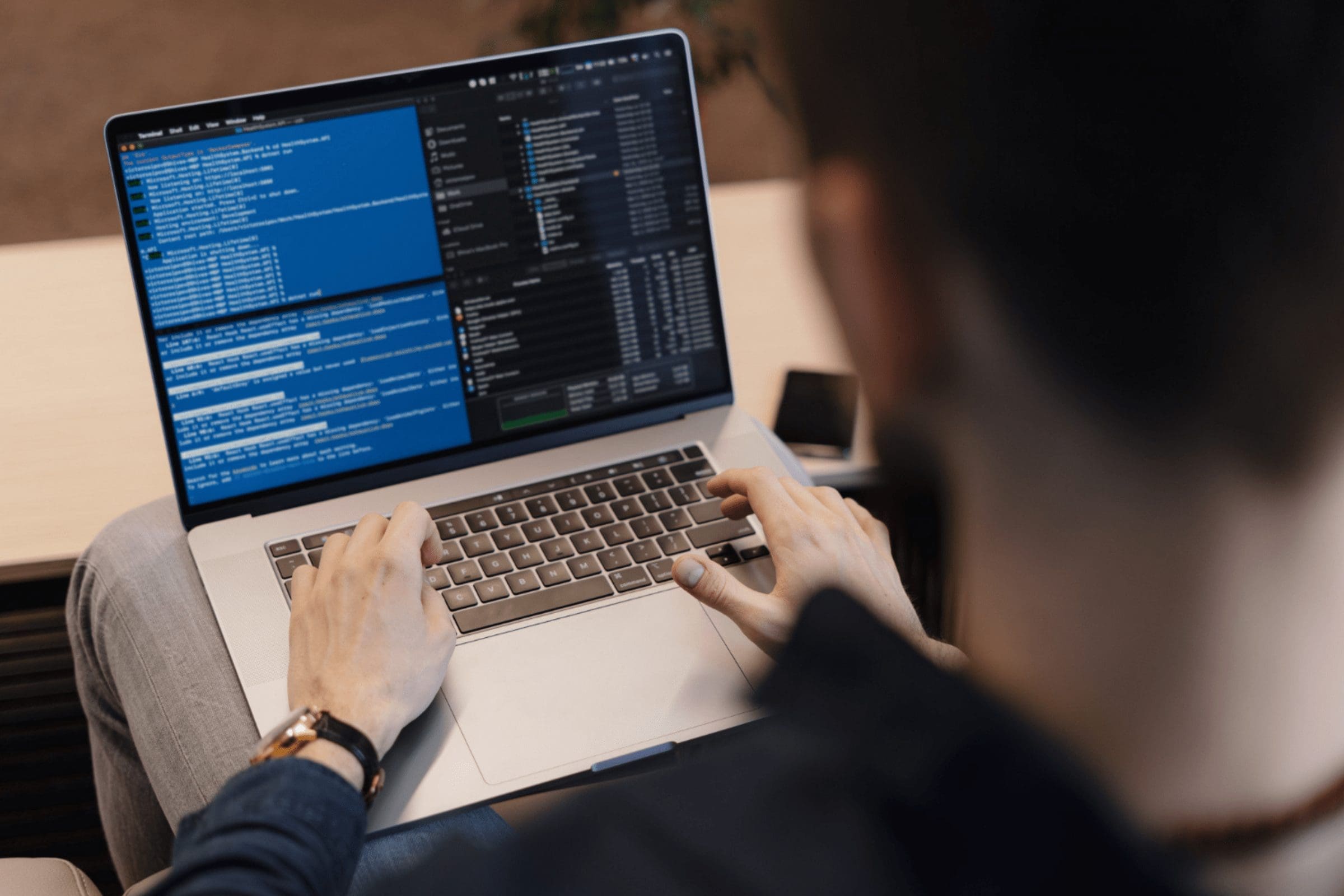 A person is working on a laptop, typing code. The screen displays multiple terminal windows and code editors with lines of text and code. The person is wearing a dark shirt and a watch, perhaps reviewing documentation for an employer of record in Mexico. The background is blurred, focusing on the laptop and the hands of the user. CodersLink 2024.