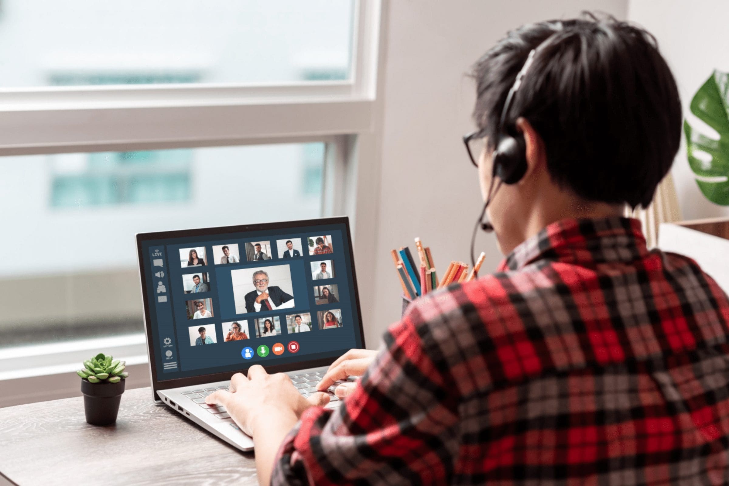 A Latin American developer wearing a red checked shirt and a headset is seated at a desk in front of a laptop, participating in a video conference. The laptop screen displays several participants. The desk features a small plant and a container with colorful pens. CodersLink 2024.