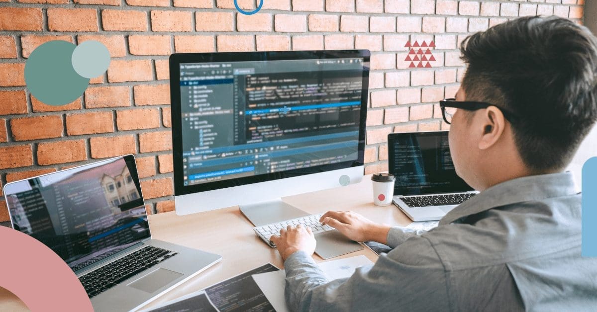 A person wearing glasses is working at a desk with three monitors displaying code. The person is typing on a keyboard in front of a large monitor. Two laptops are open on either side, also showing code. A coffee cup sits beside the computer on a brick wall background, showcasing the environment for hiring remote developers. CodersLink 2024.