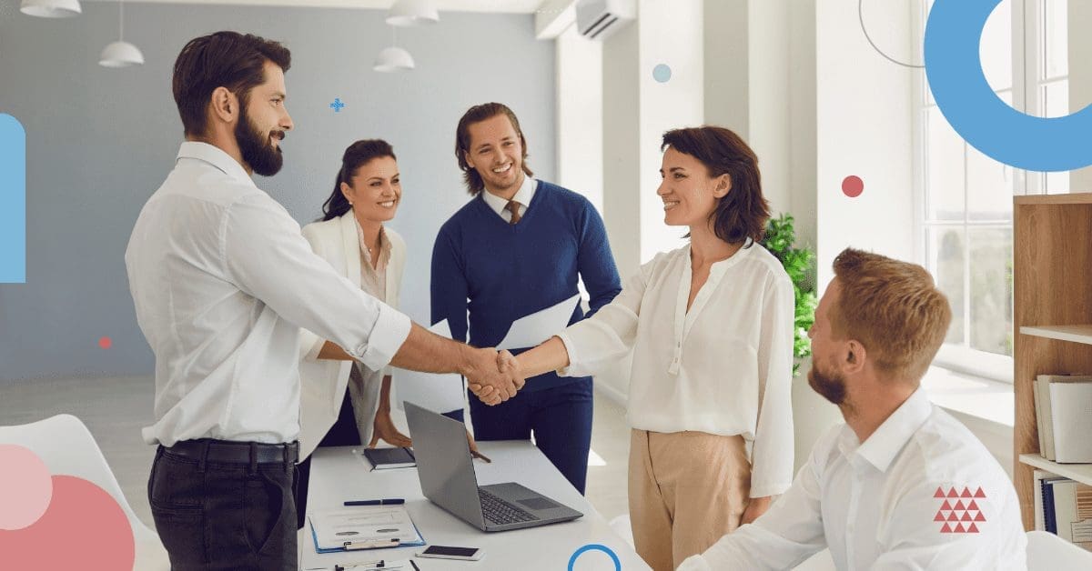 Five people in professional attire are in an office. Two are shaking hands in the center, while three others smile and observe. A laptop and documents are on the table, hinting at a collaborative discussion about hiring in Mexico. The atmosphere seems collaborative and positive. CodersLink 2024.