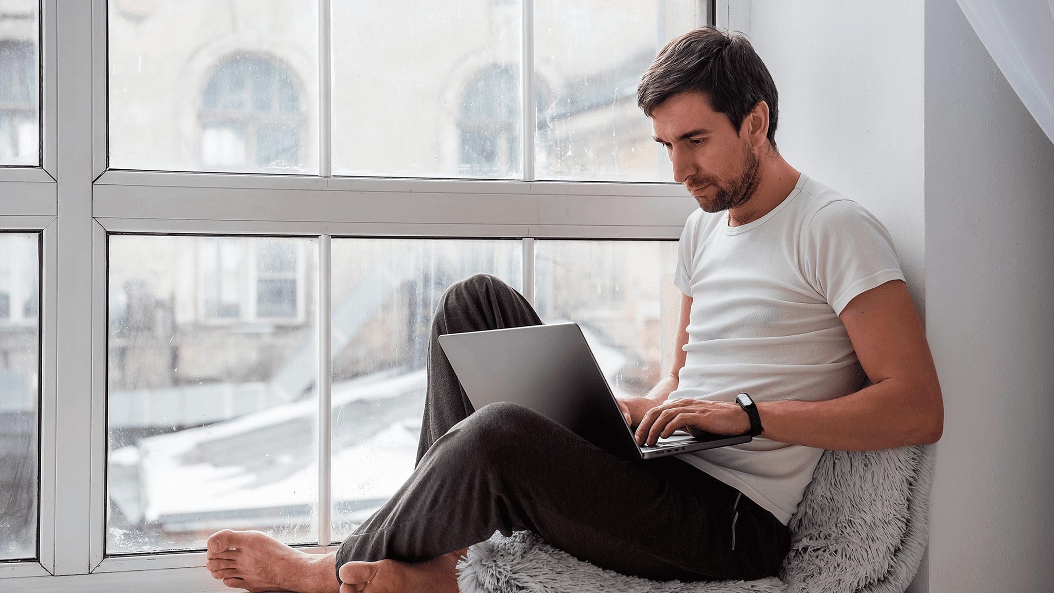 A man with short dark hair and a beard is sitting on a cushioned seat by a large window, wearing a white t-shirt and dark sweatpants. He is focused on typing on a laptop resting on his knees, delving into Mexico's Tech ecosystem. The background shows an out-of-focus view of buildings. CodersLink 2024.