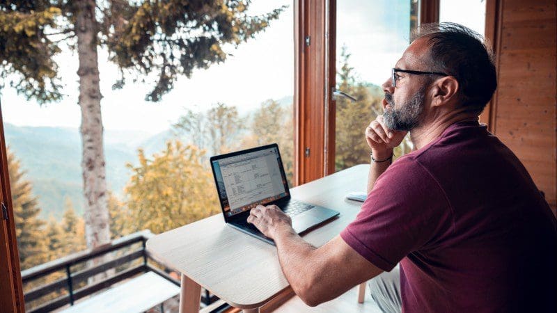 A man with a beard wearing glasses, a maroon t-shirt, and shorts is sitting at a table by a large window, working on his laptop. The background shows trees and mountains, suggesting he has reubicarse to enjoy this tranquil location while embracing trabajo remoto. CodersLink 2024.