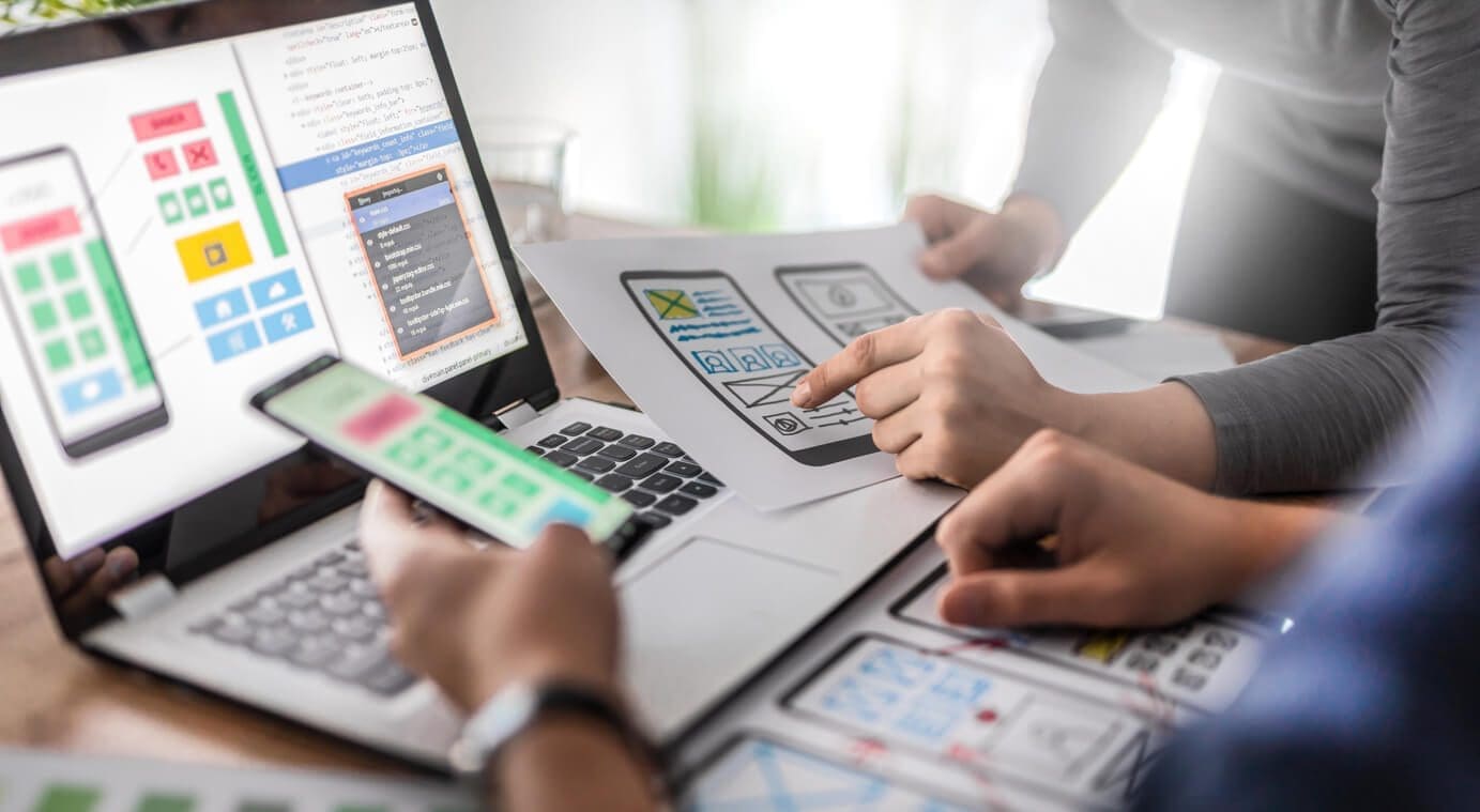 A group of people working at a desk with laptops, a smartphone, and design prints. They are engaged in a user interface design project for aplicaciones móviles, discussing and pointing at the paper designs and screen mock-ups on the devices. CodersLink 2024.