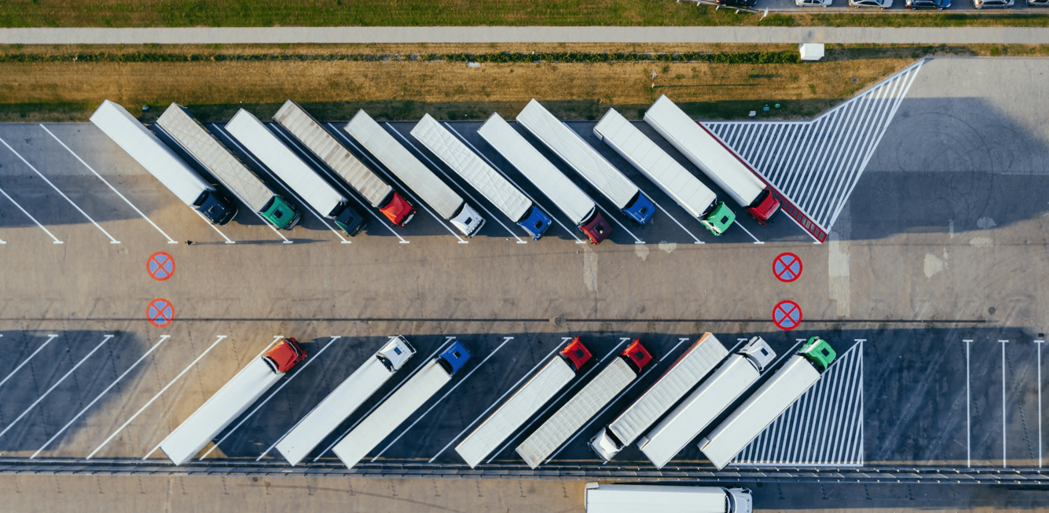 Aerial view of several trucks with trailers diagonally parked in a lot. The lot has designated parking spaces with some marked with red Xs, indicating no parking. The paved area includes green manicured grass and a row of parked cars at the top edge, showcasing a customized solution for efficient space management. CodersLink 2024.