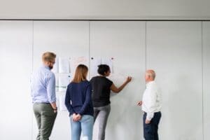 Four people are standing in front of a whiteboard. One person is drawing on the board while the others observe. The whiteboard has several papers pinned to it, likely frameworks for their Recruitment Process Outsourcing strategies. They appear to be in a discussion or collaborative brainstorming session. CodersLink 2024.