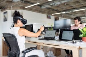 A person wearing a white outfit and virtual reality headset is seated at a desk in an office, using a pen to interact with an object in VR—demonstrating innovative tech recruitment solutions. In the background, another person is working on a laptop. Multiple computer screens and a potted plant are visible on the desks. CodersLink 2024.