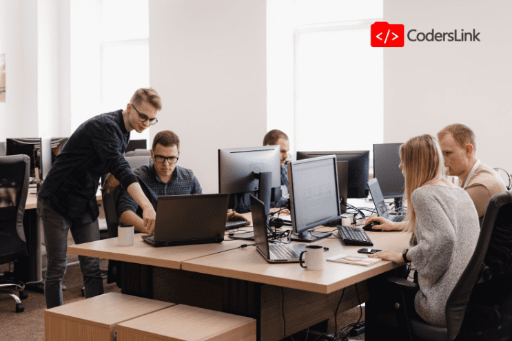 In an office setting, a group of five people collaborates on different levels around desks equipped with computers. One person points at a laptop screen while others observe. A "CodersLink" logo sits in the top-right corner, surrounded by cups and various office supplies. CodersLink 2024.