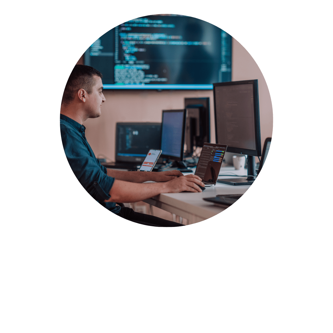 A man is sitting at a desk in a tech office environment with multiple computer monitors displaying code. Strategically, he manages his tasks while holding a smartphone and a laptop, efficiently navigating his workload to maximize productivity. CodersLink 2024.